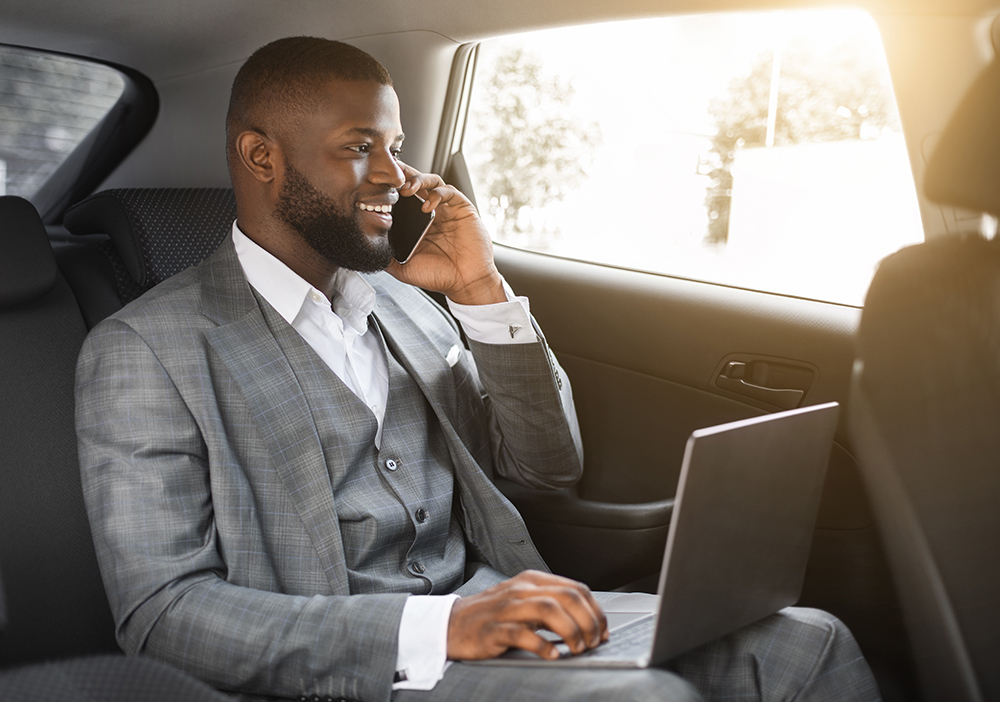 Happy black businessman talking on mobile phone and looking through window, using laptop, going to business meeting by car, african manager sitting on back seat in car, having conversation on phone